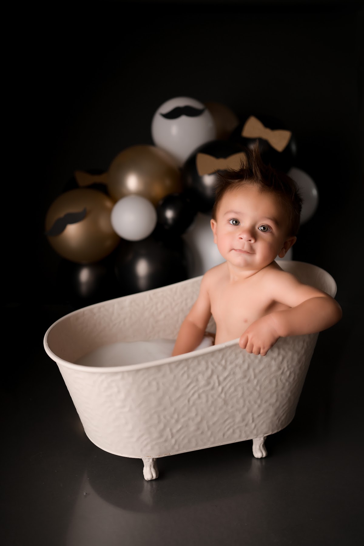 A baby boy in a bow tie at his first-birthday milk-bath session with a balloon garland