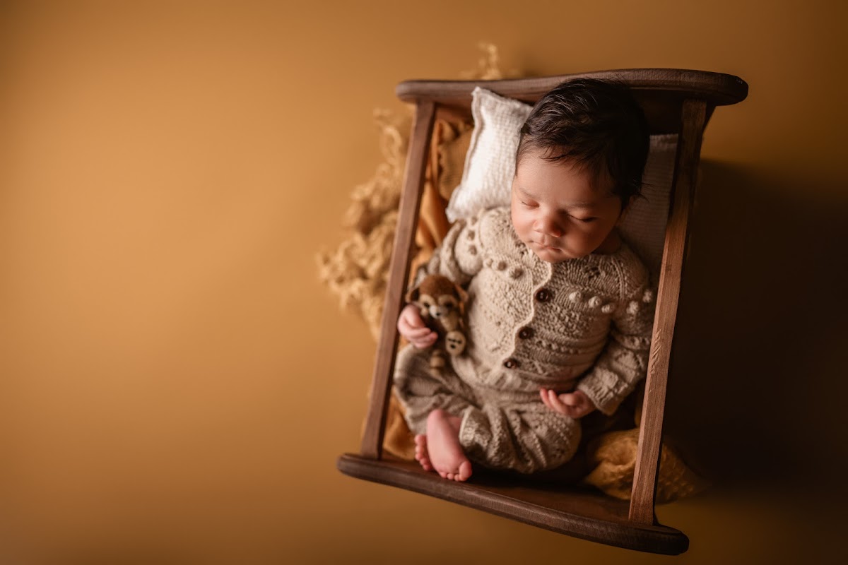A newborn in a knit outfit asleep in a tiny wooden bed in warm golden tones