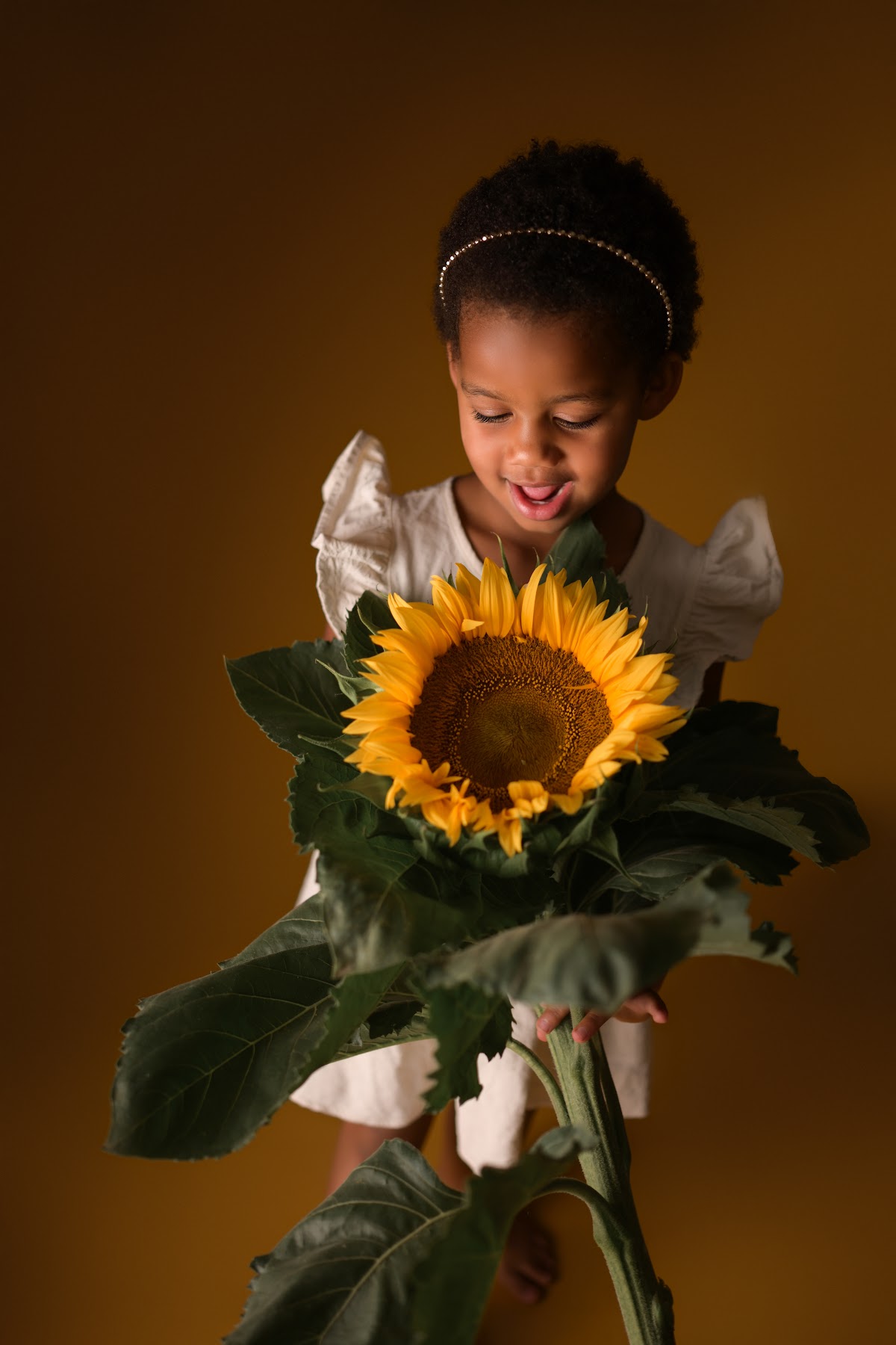 A little girl smiling as she holds a giant sunflower against a warm backdrop
